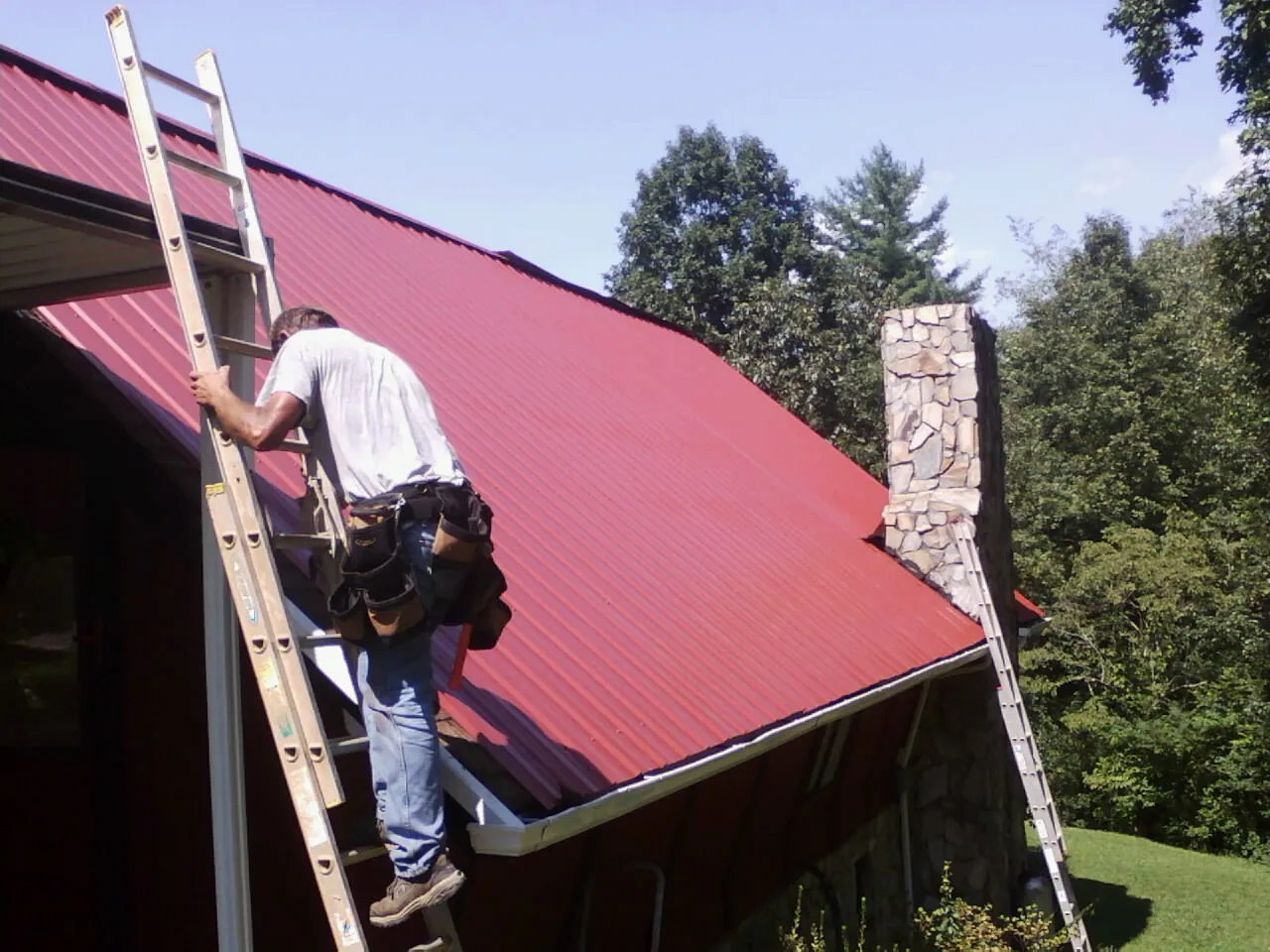 Roofer on a ladder beside a red metal roof and stone chimney — Soffit & Fascia Repair by Roofing Brothers in Black Earth
